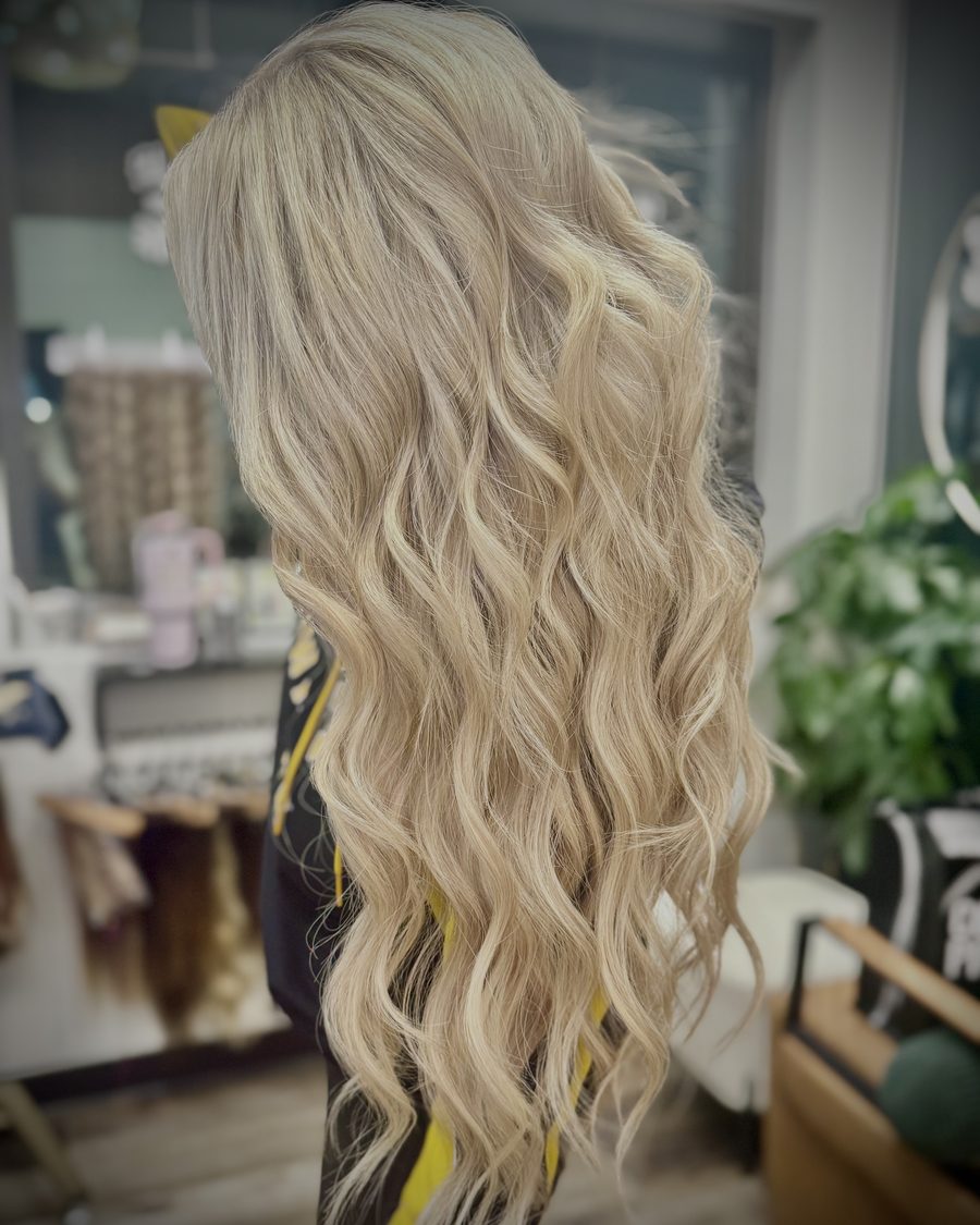 Waist-length beachy blonde waves from hand-tied IBE extensions, viewed from the side at Emerald Salon & Suites in Hiawatha, Iowa